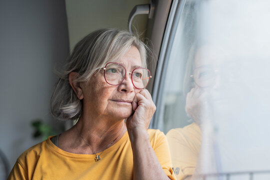 An elderly woman with grey hair and glasses gazes out the window, resting her chin on her hand. Soft natural light highlights her thoughtful expression and quiet reflective mood at home.
