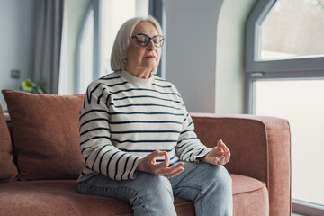 Obraz premium Older woman with closed eyes practices seated meditation and deep breathing on a couch at home, conveying calm, mindfulness, relaxation and mental wellbeing in a sunlit living room.