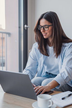 Young woman sits at a table by a window, smiling as she types on a laptop. Casual remote work setup with notebook, coffee cup and natural light conveying focus, comfort and productivity.