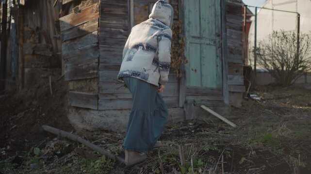 Woman walking past rustic home toward shed, wearing hooded jacket and long skirt, slow measured steps, pale winter light, neglected garden and leaning fence, gate and path underfoot, domestic chores