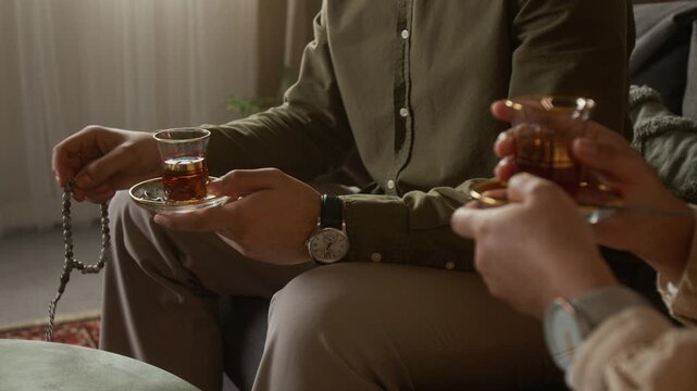 Close up shot of unrecognizable Muslim couple drinking tea together at home with focus on hands holding traditional tea glasses and praying beads, copy space