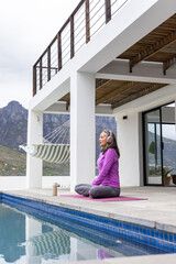 Fototapeta premium Senior woman sitting cross-legged on yoga mat at pool patio wearing purple top, headset, copy space