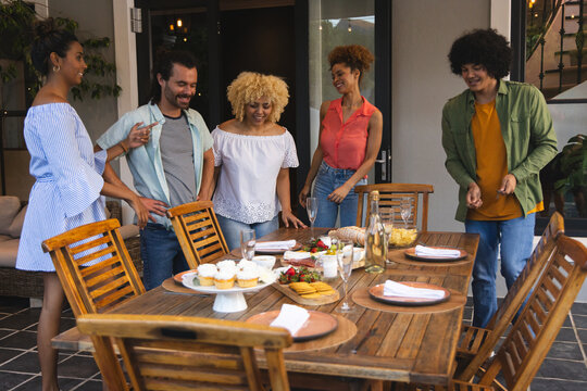 Diverse friends gathering around wooden dining table on covered patio, sharing cupcakes and wine
