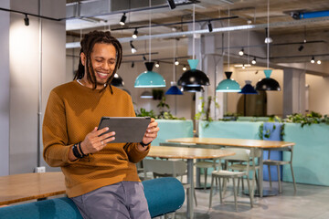 Fototapeta premium African American man in 30s wearing rust sweater leaning on teal bench using tablet in workspace