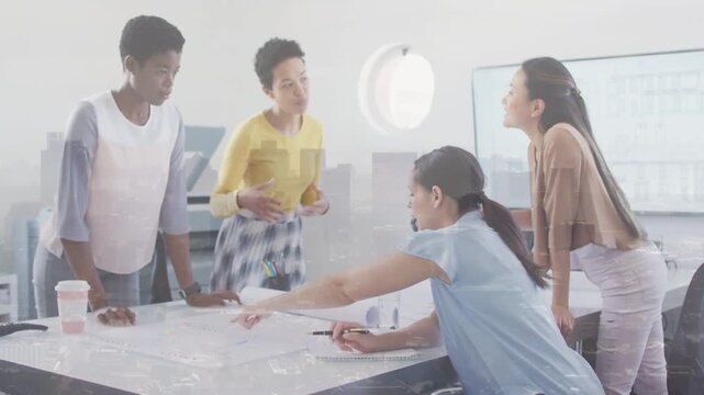 Reviewing four women wearing yellow pale blue beige tops leaning over table at office marking plans