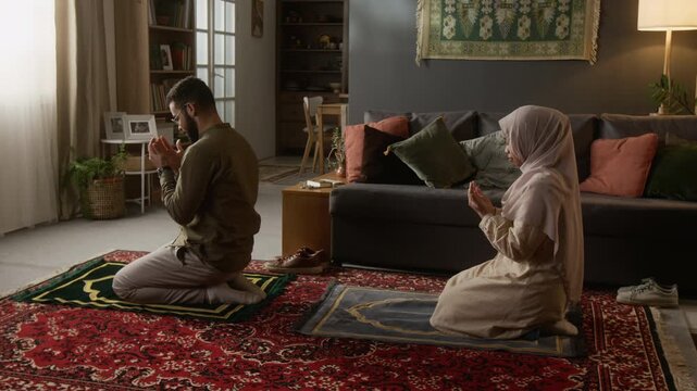 Side view shot of young Muslim couple praying together in daily religious ritual sitting on prayer mats in modern home interior with traditional elements, copy space