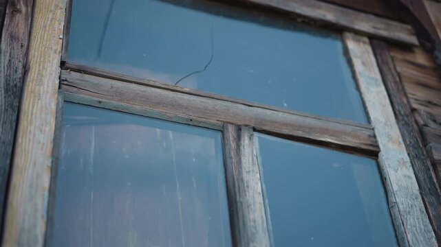 Closeup weathered wooden window with cracked glass, peeling paint, frost streaks on inner pane, aged frame of rural home, dried vine clusters at sill, soft cold light, quiet abandoned atmosphere.