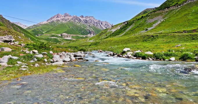 Crystal clear mountain stream flowing through green valley under blue sky in Xinjiang, China.