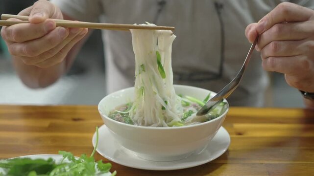 Vietnamese Noodle Closeup. Diner Pulls Noodles To Show Texture. Steaming Broth And Herbs Beneath Noodles Demonstrated. Closeup Of Diner Lifting Slippery Rice Noodles With Chopsticks