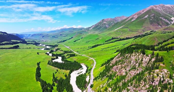 Aerial view of lush green mountain valley with winding river and road under blue sky in Xinjiang, China.