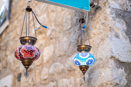 Two hanging mosaic lanterns with red and blue glass patterns on chains against a rough stone wall, showing ornate metal caps, wiring, and colorful decorative lights in a warm outdoor tourist alley