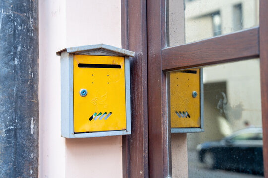 Yellow metal mailbox mounted on an exterior wall beside a wooden framed window, reflected in the glass, showing weathered paint, keyhole, and mail slot on city facade daylight with muted reflections