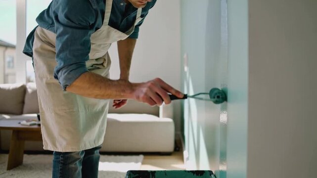 Placing roller, man wearing denim shirt, canvas apron, rolling paint down living room wall for DIY