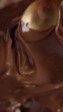 Close-up of melted chocolate being spread over hazelnuts with a spoon. Smooth glossy texture and dessert preparation process.