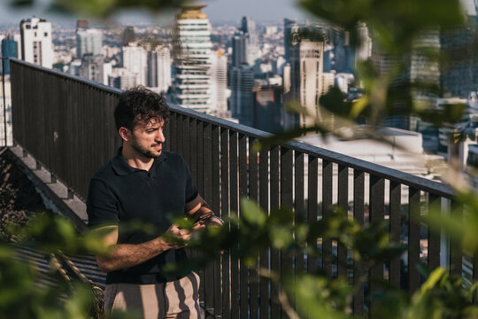 Young man with curly hair standing on a high-rise rooftop, holding a smartphone and looking at the urban cityscape. Bangkok, Thailand