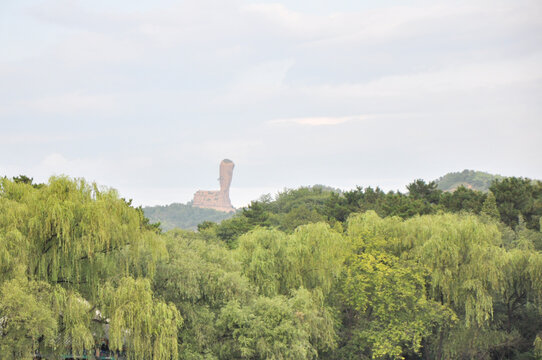 A distant view of the iconic Bangchui Mountain (Qingchui Peak), a unique reddish rock formation shaped like a pestle, rising above a sea of lush green willow and pine trees under an overcast sky