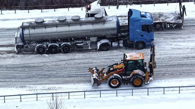 Truck struggling on icy winter road after snow, backhoe assisting clearance, aerial view during blizzard
