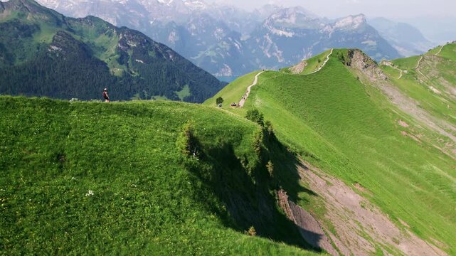 Aerial view of hikers walking along the green Stoos Ridge in Switzerland, with sweeping alpine peaks and dramatic mountain scenery in the background.