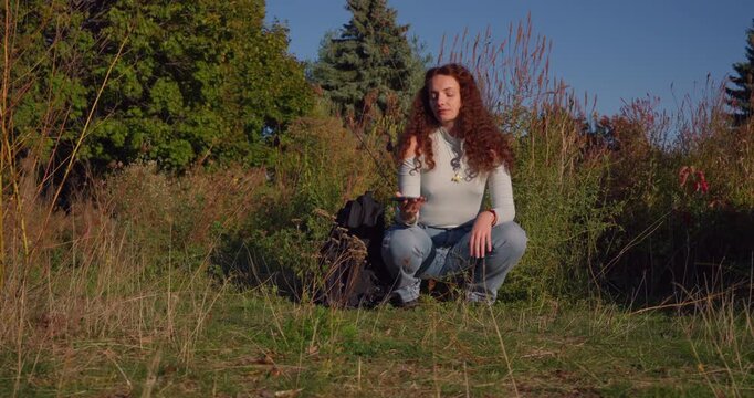 A young woman crouches in sunlit grass, turning her phone in her hands as she pauses, reflects, and questions the place screen time takes in everyday life.