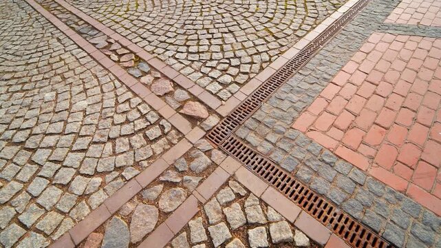 Close view of worn cobblestone paving with brick inlays and a metal drainage channel, showing urban texture, street design, and pedestrian walkway detail.
