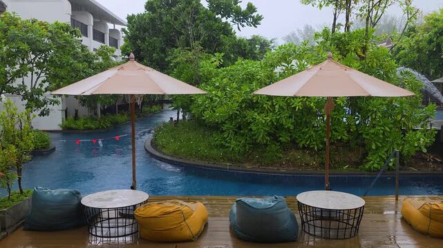 Wet wooden deck with colorful bean bag chairs and patio umbrellas overlooks curved pool surrounded by lush tropical garden during monsoon rain at JW Marriott Khao Lak Resort