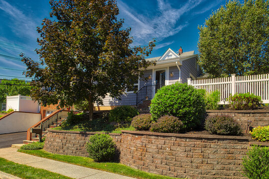 Bright exterior of a single-family home featuring a tiered masonry retaining wall, manicured shrubs, and mature trees. 