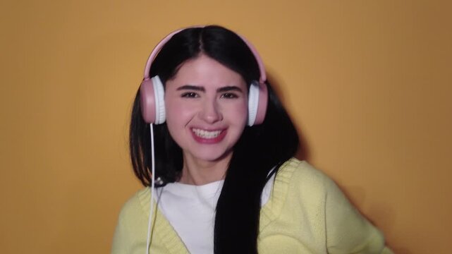 Cheerful young brunette woman wearing headphones listening to her favorite song, dancing energetically and making funny faces on a solid yellow background, enjoying the music and smiling at camera