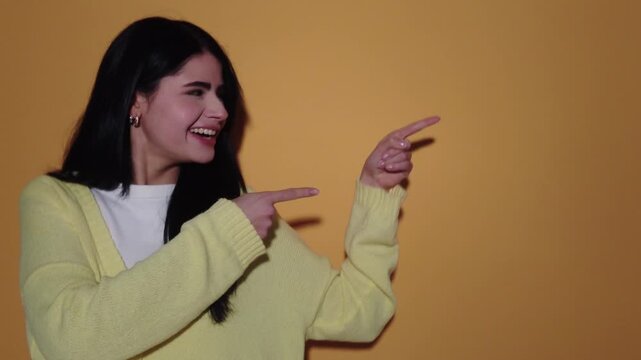 Happy young woman pointing her fingers to the side with excitement, smiling and dancing while presenting a blank space for advertising copy on an isolated yellow studio background