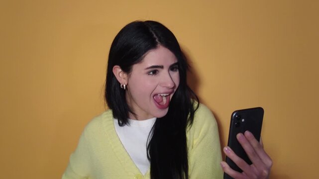 Cheerful young woman with long dark hair holding her smartphone, displaying a wide range of emotions from angry to happy and excited while having a video call on a yellow background