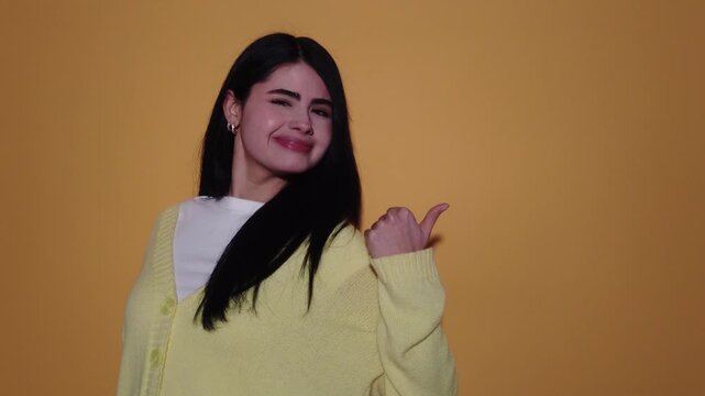 Cheerful beautiful woman with dark hair pointing her finger to the side, smiling and showing various positive emotions while presenting an empty space on a yellow background in a studio