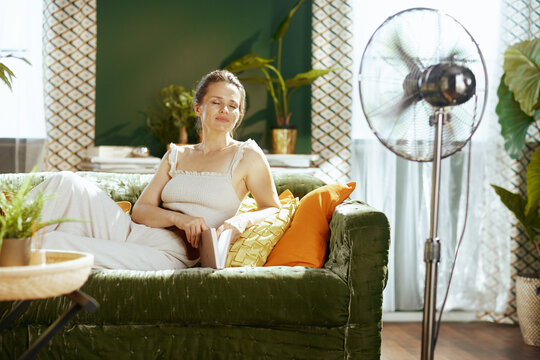 A middle-aged woman, eyes closed in serenity, relaxes on a green velvet couch with a book, enjoying the cooling breeze from a standing fan in her bright, plant-filled living room.