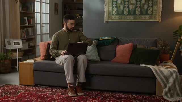 Full length shot of young Muslim man starting daily prayer at home, checking watch, taking off shoes aying out prayer rug on floor in cozy home setting with traditional elements, copy space