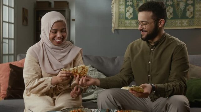 Tilt up shot of Muslim young couple enjoying traditional meal together sitting on couch at home and sharing flatbread with candid smiles