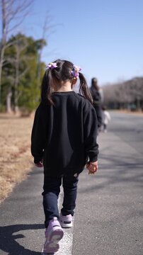 Little girl walks forward with pigtails and black hoodie. She follows adult on paved path under blue sky. Trees line the path with dry grass nearby. Child moves calmly, eyes focused ahead