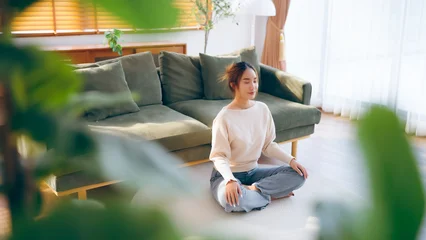 Fotobehang Lotusbloem Happy young Asian woman practicing yoga and meditation at home sitting on floor in living room in lotus position and relaxing with closed eyes. Mindful meditation and wellbeing concept  © Monster Ztudio