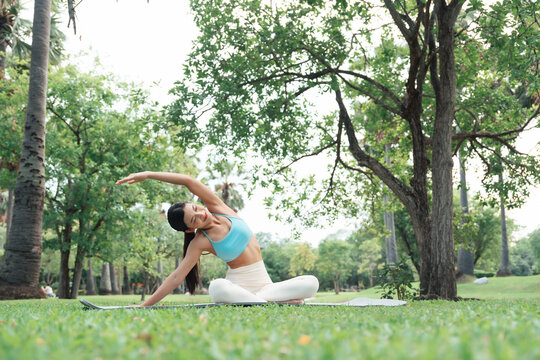 Asian woman practicing seated side stretch yoga pose outdoors in a peaceful park, wearing athletic wear. Promotes mindfulness, flexibility, and a healthy, balanced lifestyle surrounded by nature