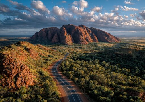 Aerial view of iconic Mount Uluru in Australia surrounded by lush vegetation and winding road under a dramatic sky with clouds at sunset