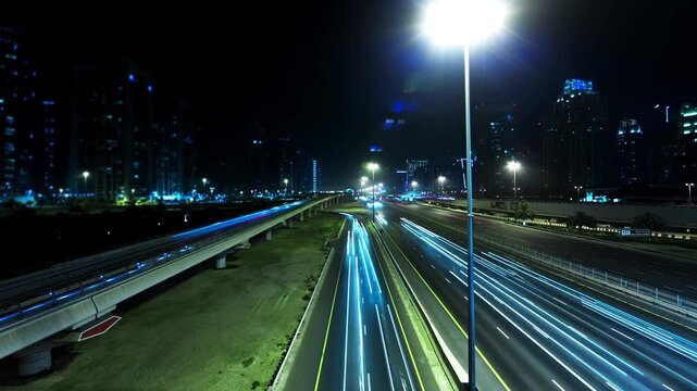 Time-lapse footage of a multi-lane highway at night, featuring dynamic light trails from fast-moving vehicles. A parallel elevated road or monorail track runs alongside the highway, while illuminated.