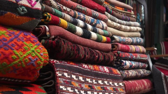 Colorful traditional kilim rugs and textiles stacked high in a bustling market stall in motion.
