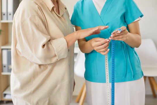 Elderly patient waist measuring by nurse holding tape and looking at scales during healthcare examination for metabolic syndrome, BMI and cardiac risk check. Senior woman on medical checkup