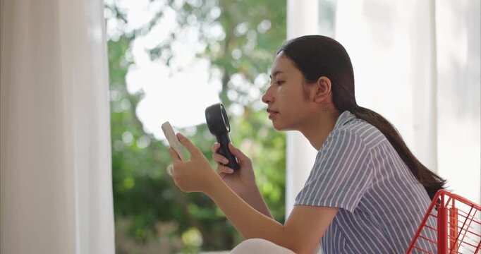 Young Asian woman using portable fan and smartphone in hot weather at home, feeling sweaty and exhausted from summer heatwave in living room.