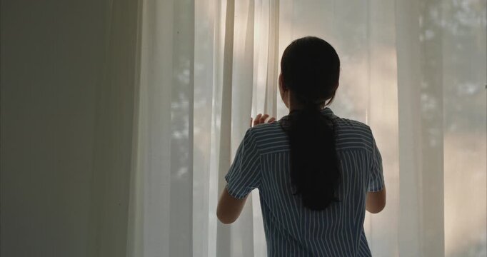 cheerful young Asian woman standing by the window and happily opening both curtains to let the soft natural morning light into her house. 