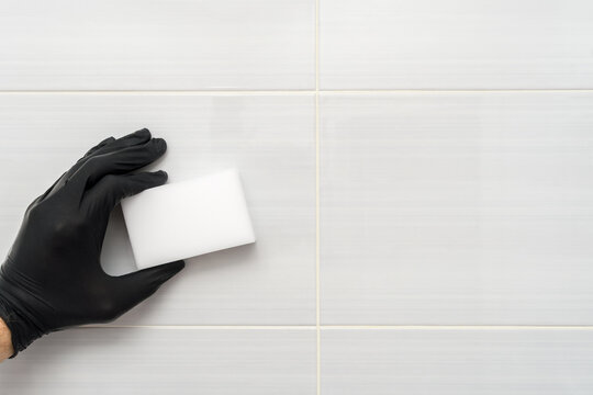 Black gloved hand holds melamine sponge near bathroom tile wall for household cleaning, hygiene, stain removal and care.
