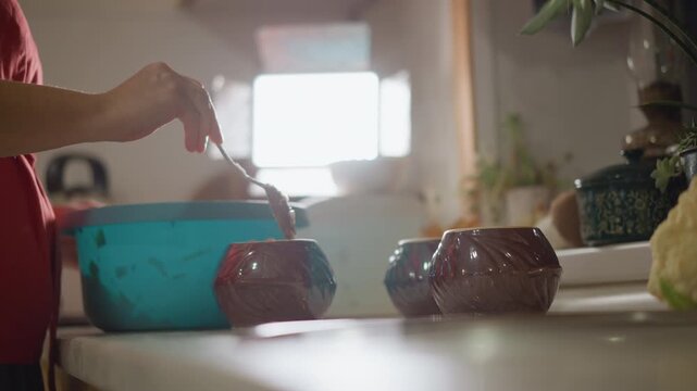 Close up hands stirring blue bowl, soft backlight on countertop, ceramic ramekins and houseplant in background, domestic prep with gentle rhythm, home cook measuring ingredients and mixing batter.