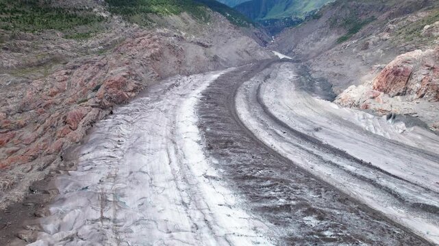 Aerial view of a melting alpine Aletsch glacier with dark medial moraines carving through ice, revealing climate change impacts in a rugged mountain valley of Switzerland via drone 4k