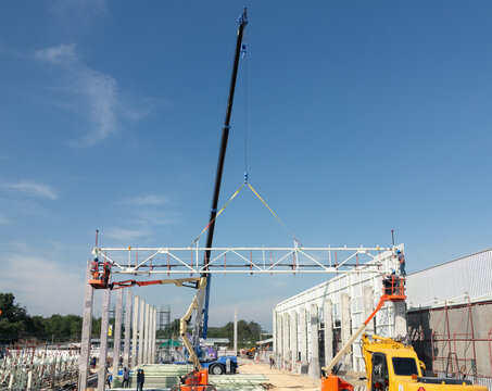 Construction crane lifts metal truss structure into position, worker on boom lift monitors alignment and placement. Represent steel construction, industrial building projects and teamwork.