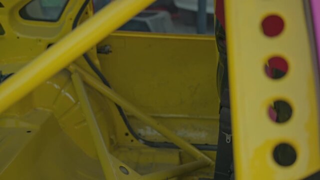 A smooth cinematic pan reveals the weathered yellow interior of a stripped-down race car, highlighting the sturdy metal roll cage and industrial textures under natural light.
