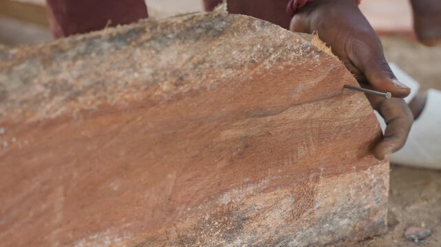 Close up black hands chiseling timber, removing rough bark and smoothing plank edge with hammer and chisel, sawdust scattering across outdoor workshop bench, focused artisan technique and rustic
