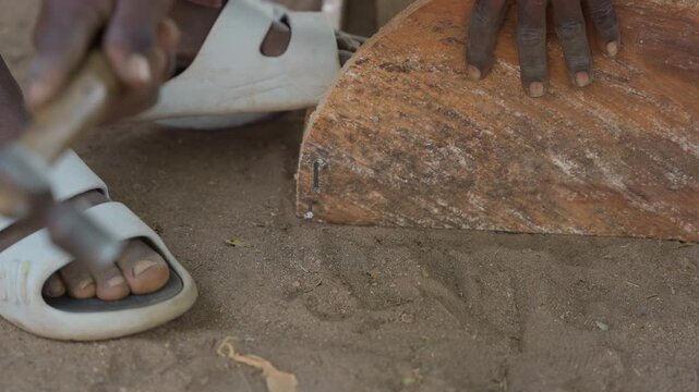 Closeup black hands hammering wood shaping rough log on dirt floor, white sandals, worn hammer, tactile shavings and grain, concentrated rustic carpentry skill in outdoor workshop