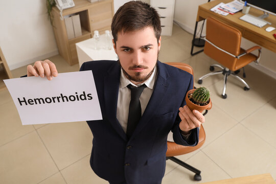 Young businessman holding paper with word HEMORRHOIDS and cactus in office
